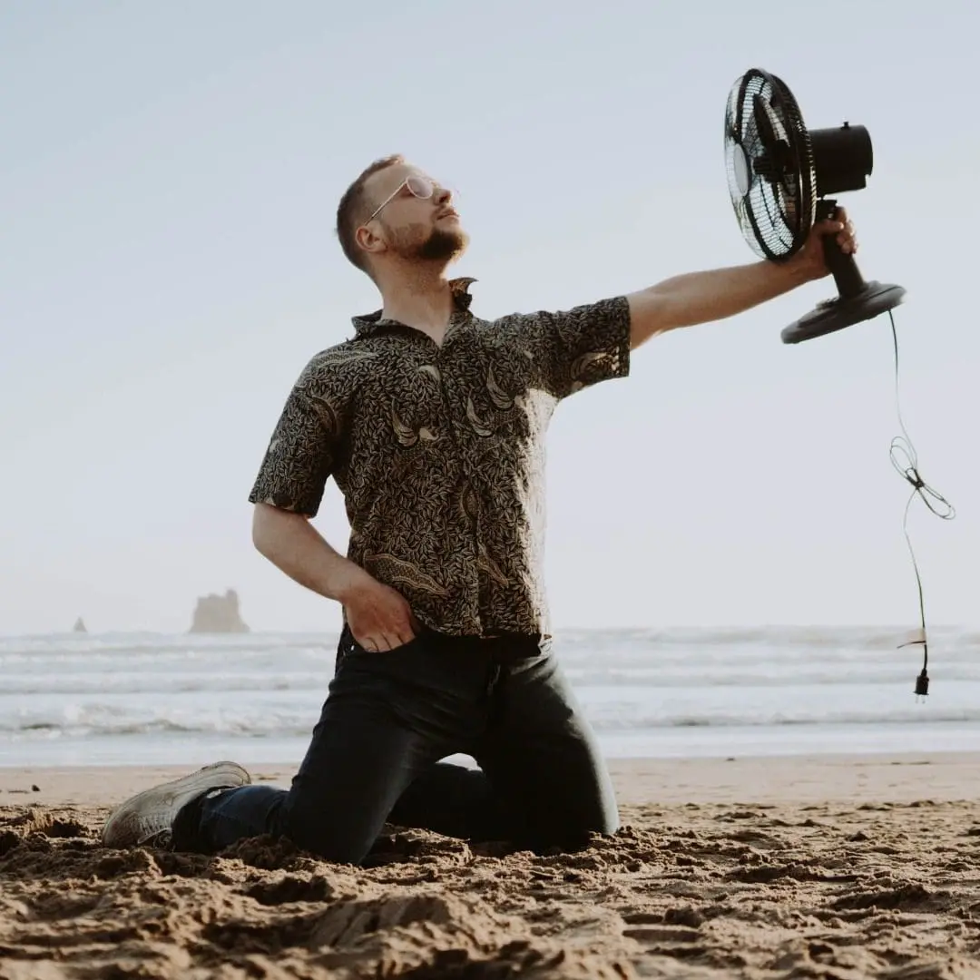 Man on the Beach Playing with an Unplugged Fan A man is playing with a fan on the beach that is not plugged in, likely pretending to use it in the breeze or as a playful prop