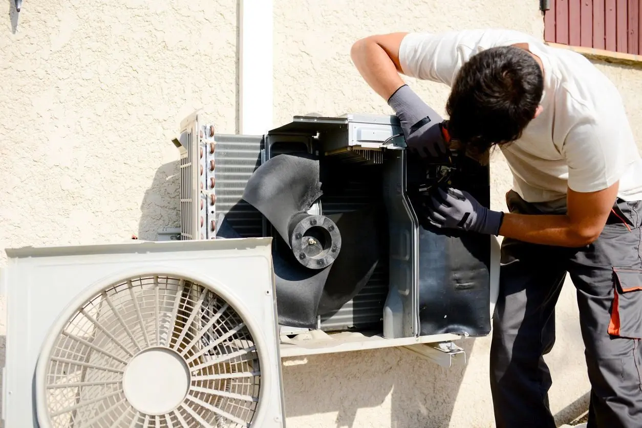 An air conditioning repair technician is working on an air conditioner outside, possibly diagnosing or fixing the unit.