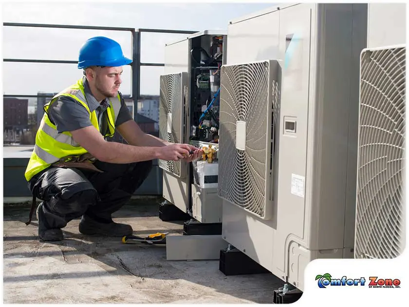 Technician in Yellow Vest and Hard Hat Fixing Air Conditioner on Roof of Residential Building A man in a yellow vest and hard hat is fixing an air conditioner on the roof of a residential building, ensuring the unit is operating properly
