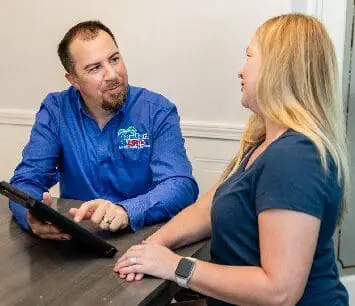An HVAC service technician is sitting at a table with a homeowner, discussing a free estimate for heating and air conditioning services, with paperwork and a tablet on the table.