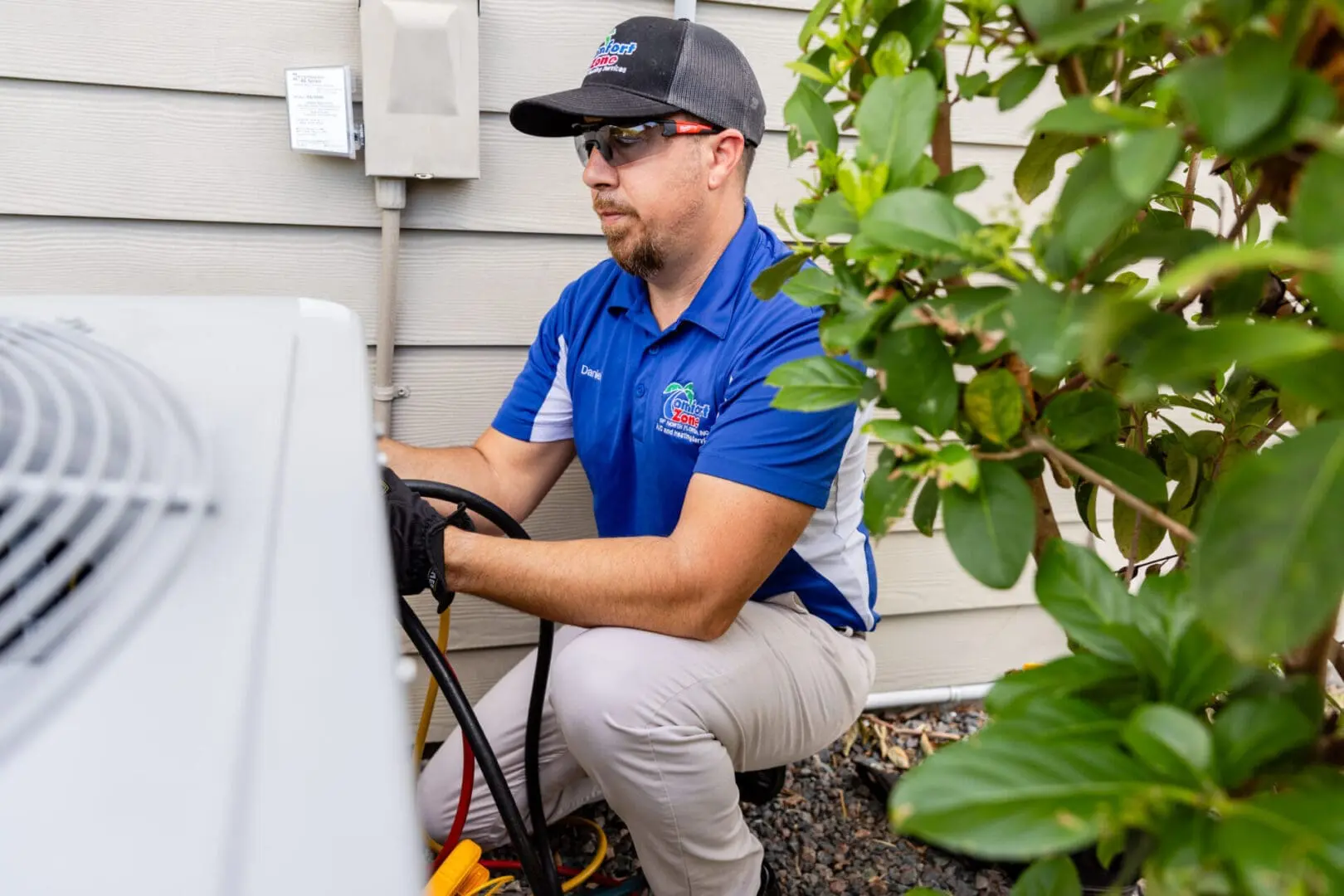 A service technician wearing a uniform and safety gloves is working on an outdoor air conditioning unit in a shaded backyard, using tools to inspect and repair the system A service technician wearing a uniform and safety gloves is working on an outdoor air conditioning unit in a shaded backyard, using tools to inspect and repair the system