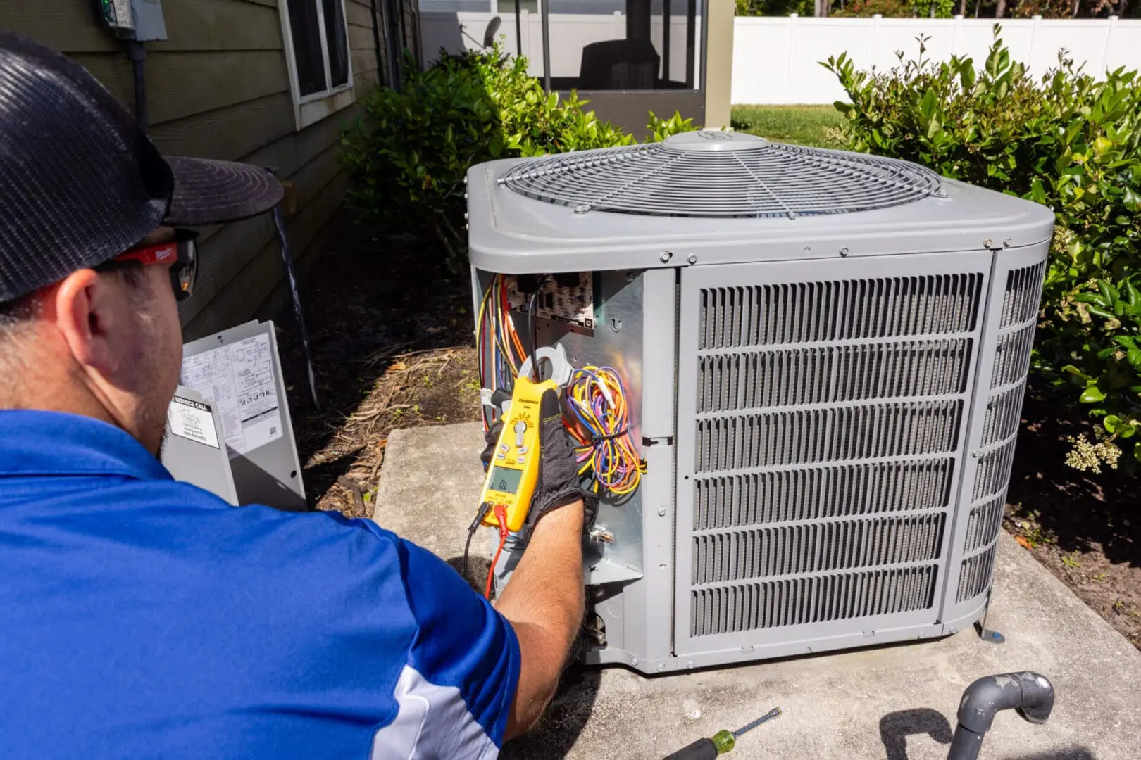 A technician wearing a uniform is working on an outdoor air conditioning unit, using tools to inspect and repair the system on a sunny day.
