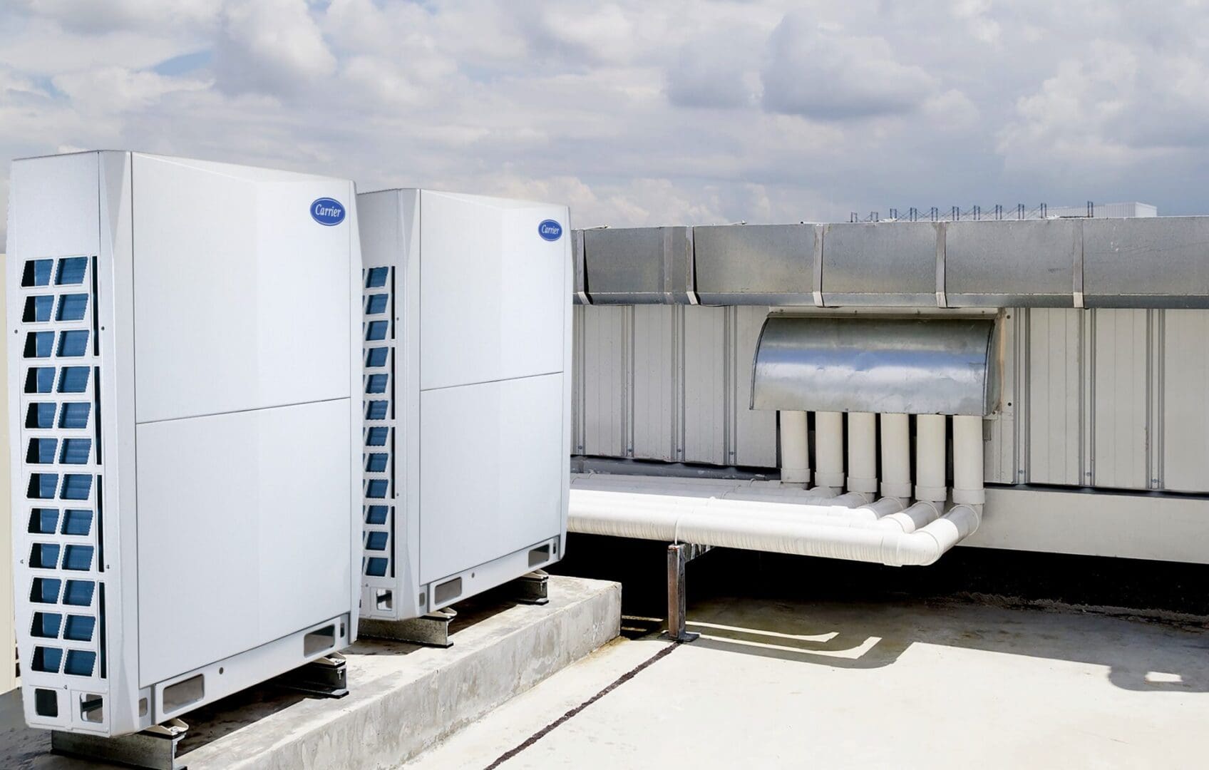 Two large industrial air conditioning units on a building rooftop, connected to ventilation ducts under a cloudy sky.