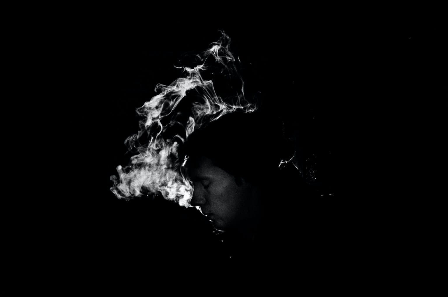 A monochrome image of a person exhaling smoke, creating a swirling pattern against a dark background.