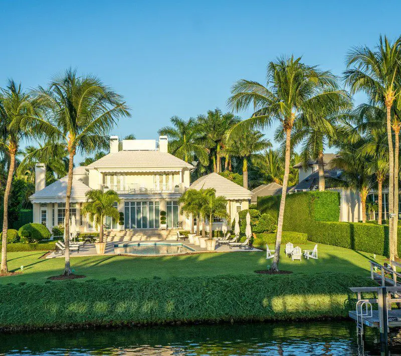 Luxurious waterfront mansion surrounded by palm trees with a well-manicured lawn and dock under a clear blue sky.