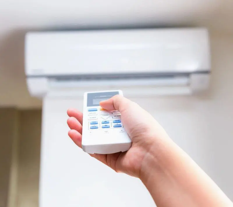 A person's hand holding a remote control aimed at an air conditioning unit on a wall.