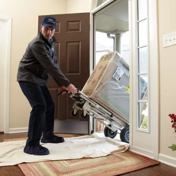 Delivery man using a hand truck to move a large boxed appliance into a house, taking care to protect the floor with a rug.