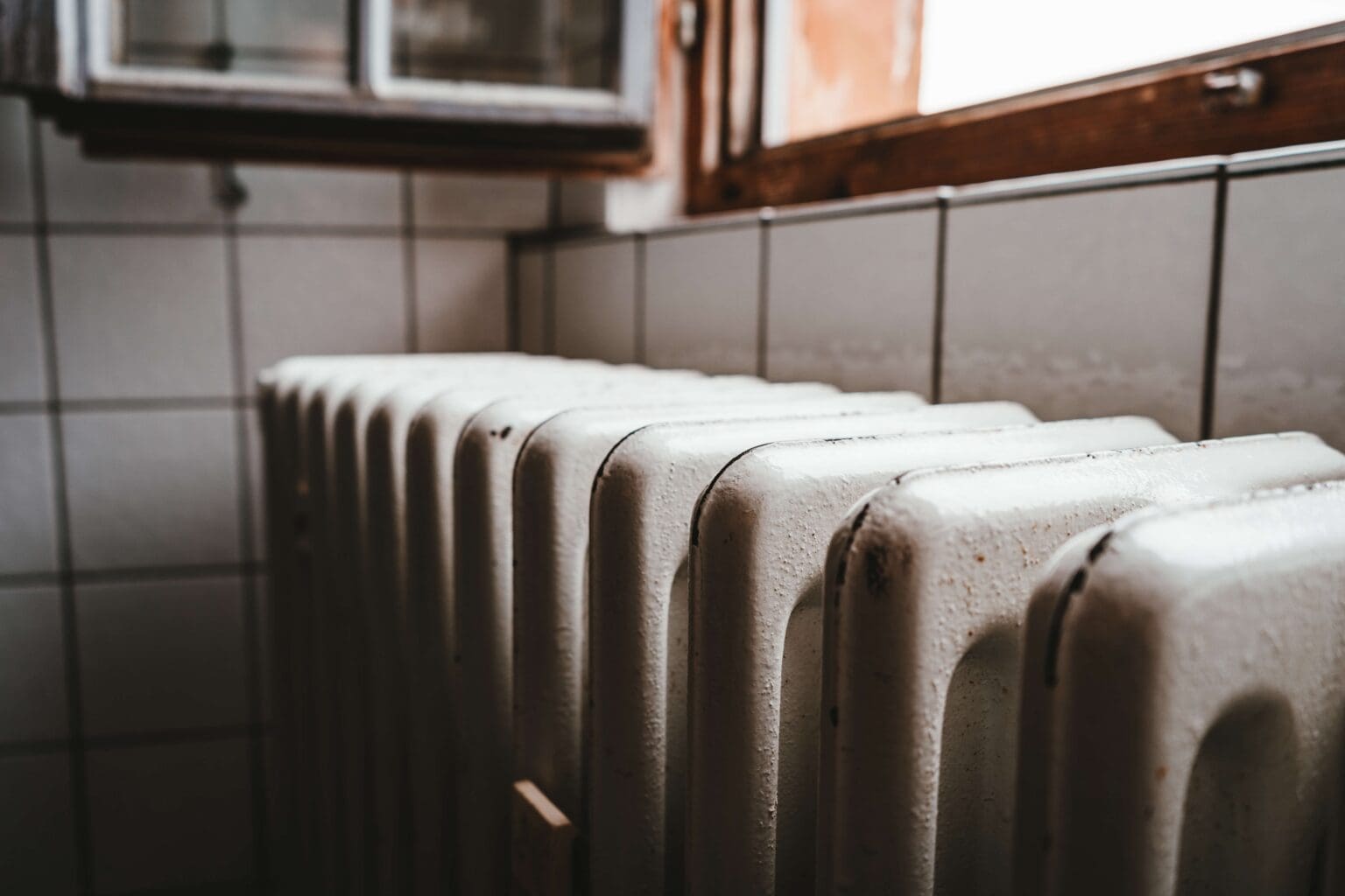 An old white radiator situated below a window in a kitchen with tiled walls, emitting warmth in a dimly lit setting.