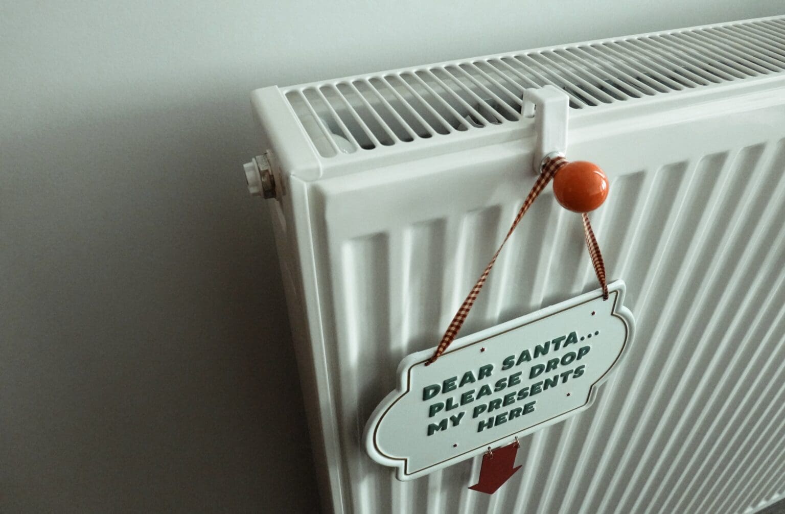 A white radiator with a decorative sign that reads "dear santa, please drop my presents here" hung by a red ribbon.
