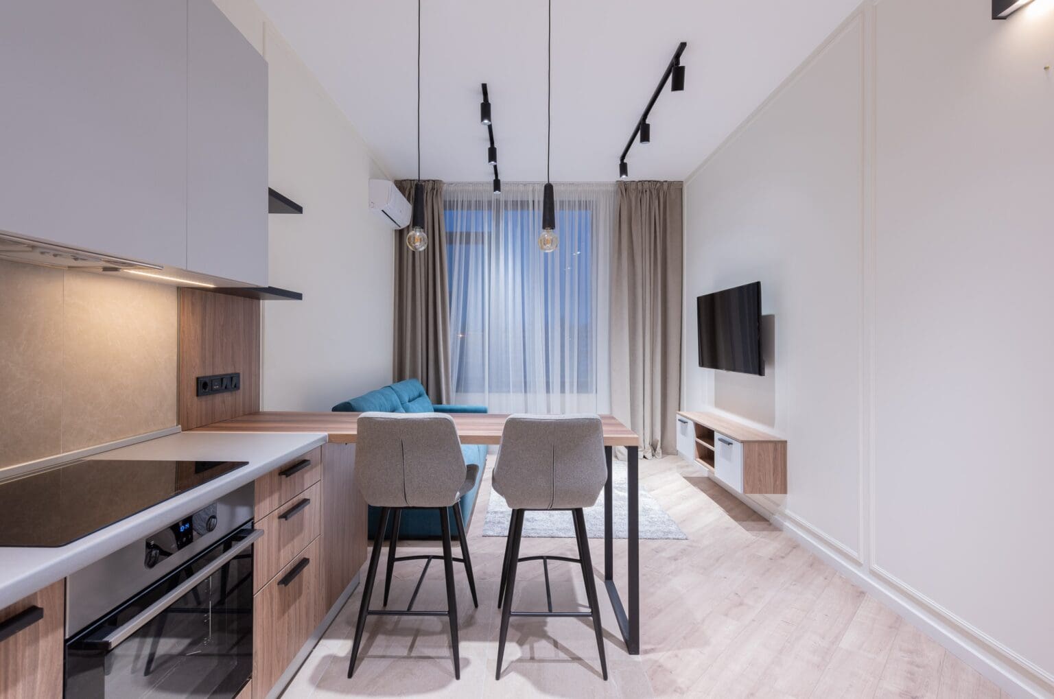 Modern kitchen with a dining area featuring a wooden table, two grey chairs, pendant lights, and integrated appliances, leading to a cozy living space with a tv.