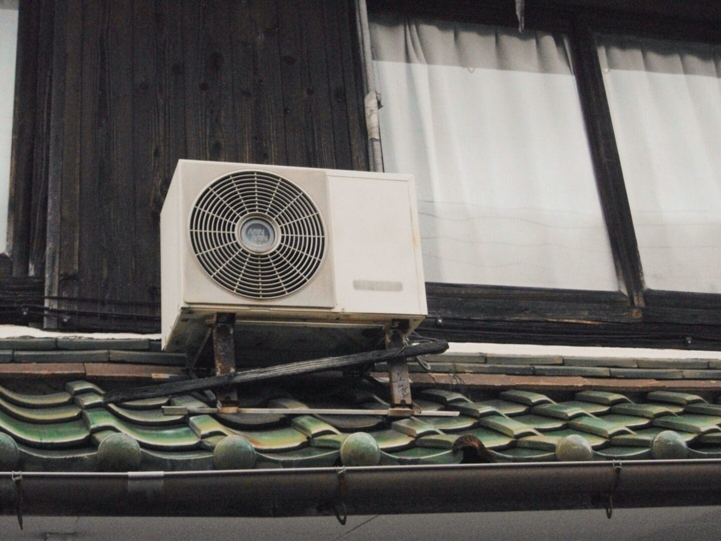 Air conditioning unit mounted on a tiled roof near a wooden window frame.