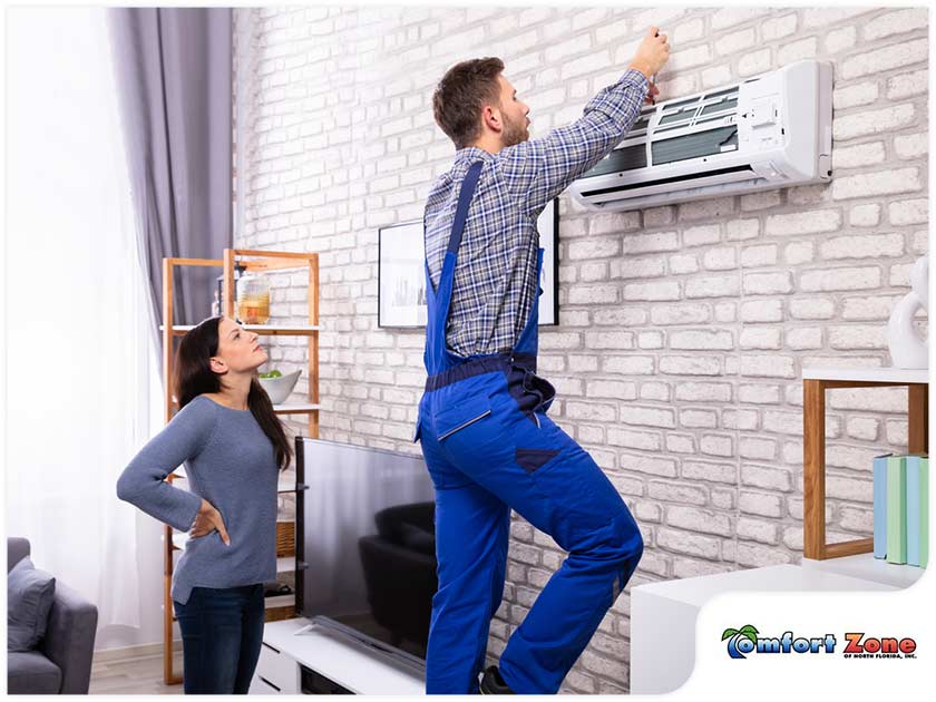 A technician in a blue uniform installs an air conditioner as a woman observes in a modern living room with brick wall.