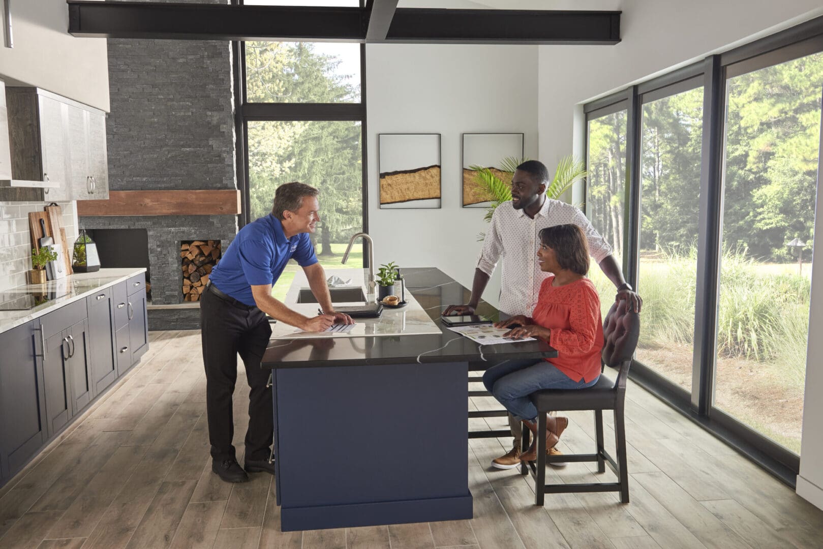 Three people discuss kitchen design plans in a modern kitchen with large windows and a central island.