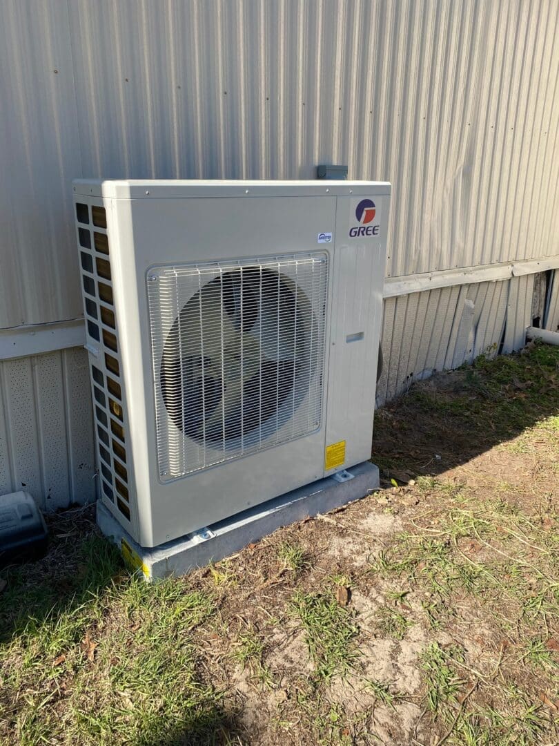 Outdoor gree air conditioning unit installed beside a building, on a grassy surface with a metal fence in the background.