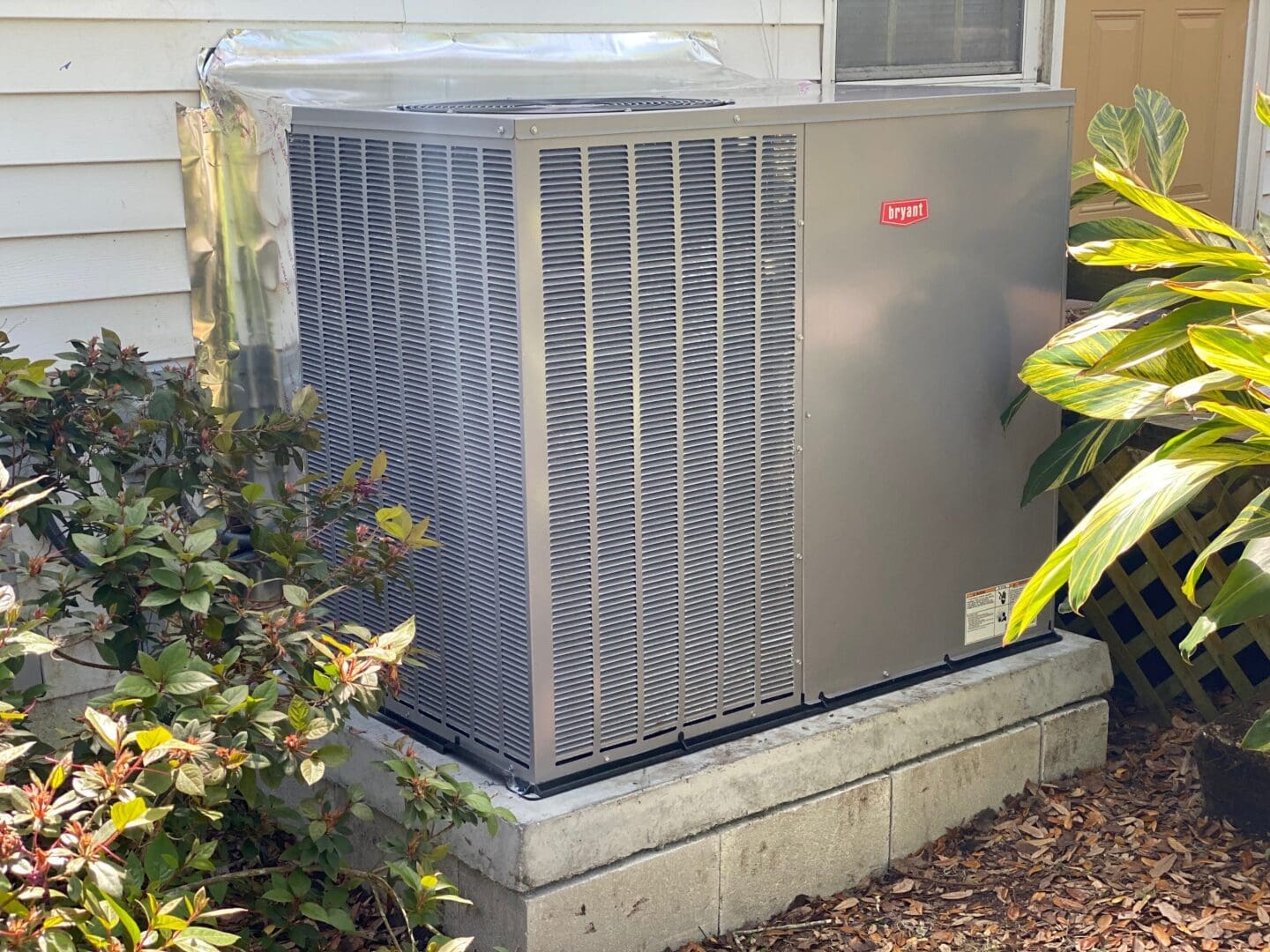An outdoor hvac unit from bryant, installed on a concrete slab next to a house, surrounded by bushes and shaded by trees.