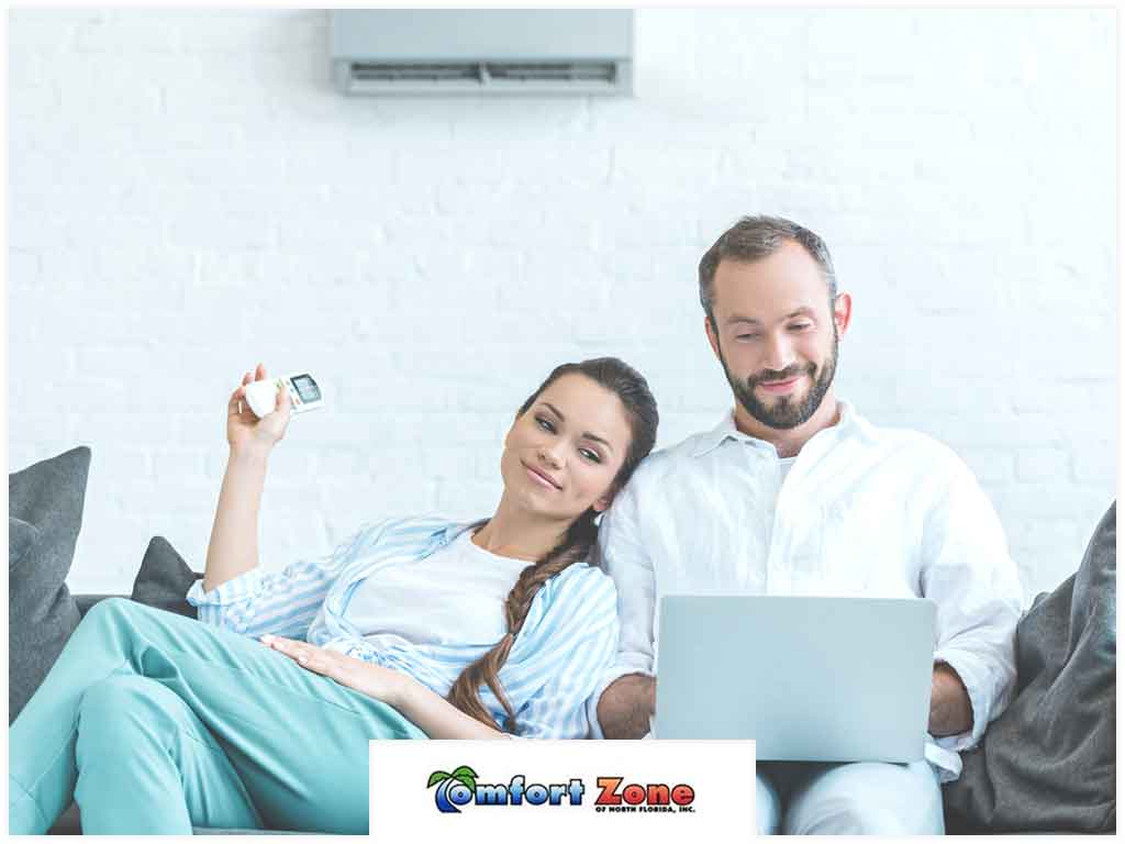 A couple relaxed on a couch, the woman holding a remote and the man using a laptop, with an air conditioner unit visible above.
