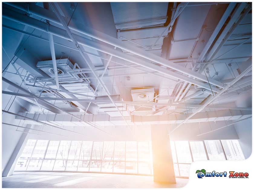 Bright, modern industrial interior showing exposed ceiling pipes and beams with large windows and a central column, with a partial logo reading "comfort zone.