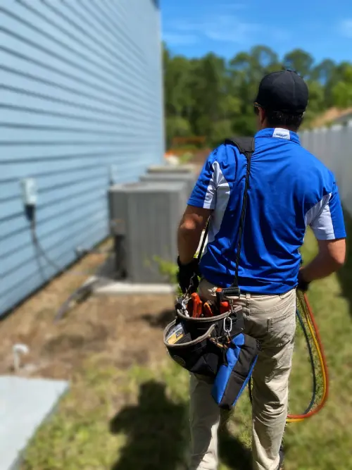 A technician in a blue shirt and khaki pants, equipped with a tool belt, looks towards an outdoor ac unit beside a house.