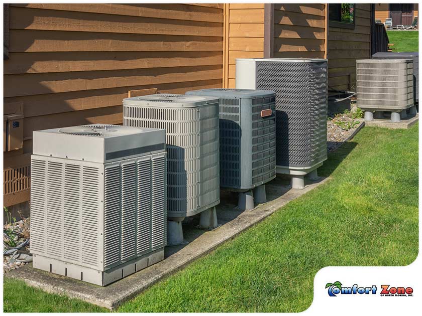 Five air conditioning units outside a residential building on a sunny day, positioned on concrete slabs with green grass and wooden siding visible.