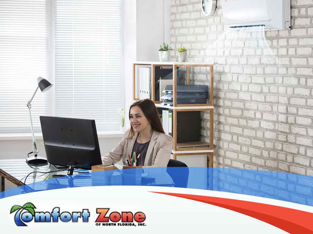 A woman smiling at a desk in a modern office with a computer, lamp, and air conditioning unit in the background.