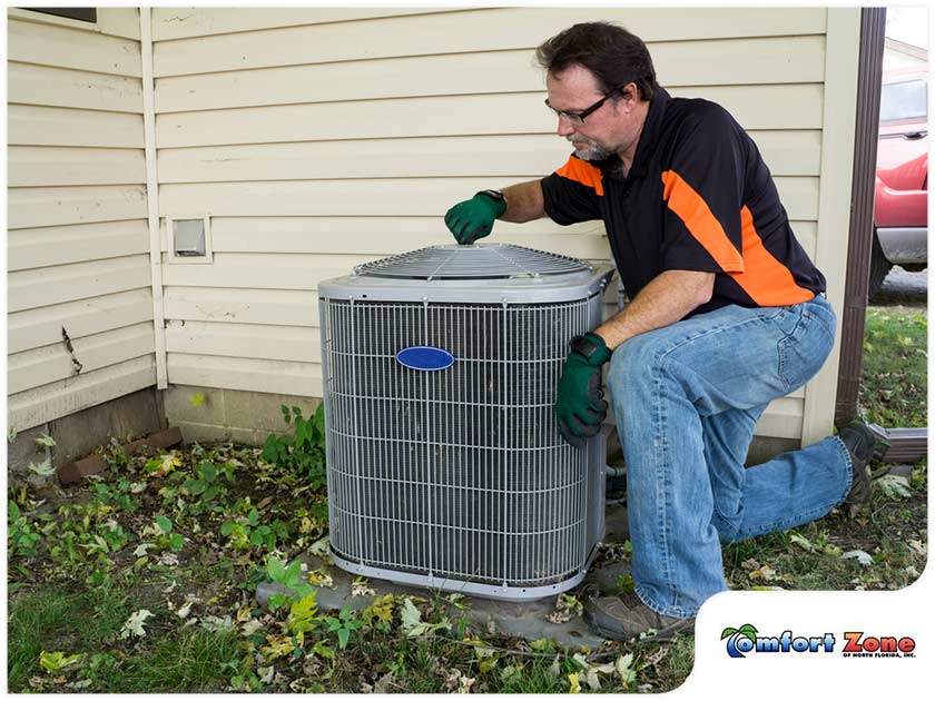 A technician in gloves and sunglasses services an outdoor air conditioning unit beside a house.