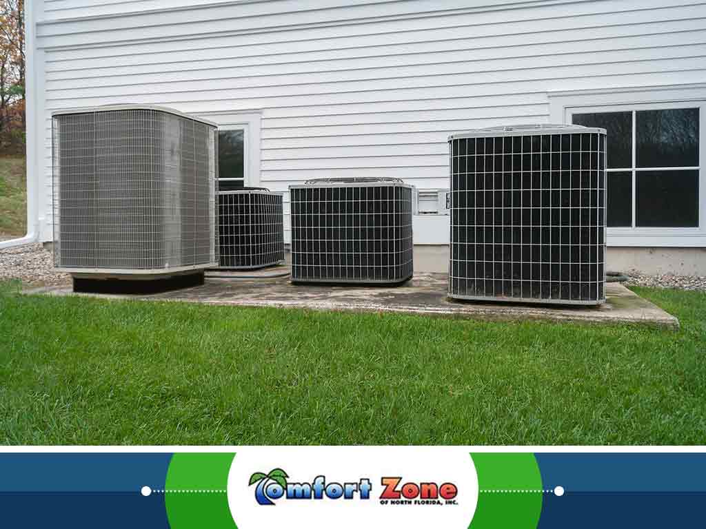 Four air conditioning units of varying sizes installed outside a white-sided building, set on a concrete slab with a grassy backdrop.