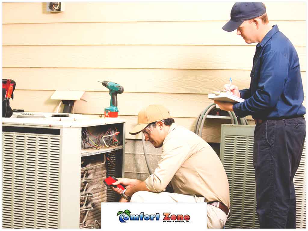 Two technicians repairing an outdoor ac unit, one working on the machinery while the other takes notes.