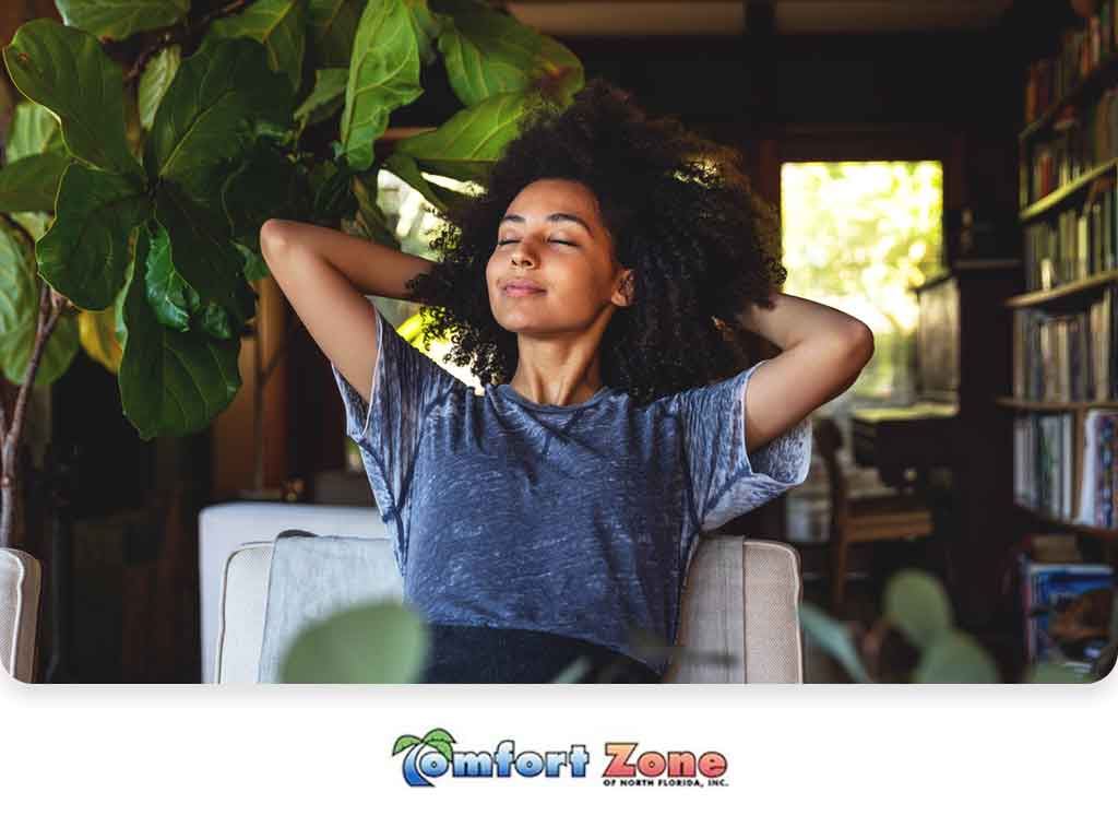 A relaxed woman sits in a chair with her hands behind her head, smiling, in a cozy room surrounded by books and plants.