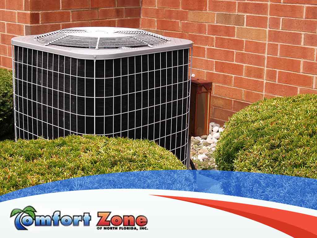 Air conditioning unit outside a brick building, surrounded by shrubbery and a logo of comfort zone of north florida, inc. visible in the foreground.