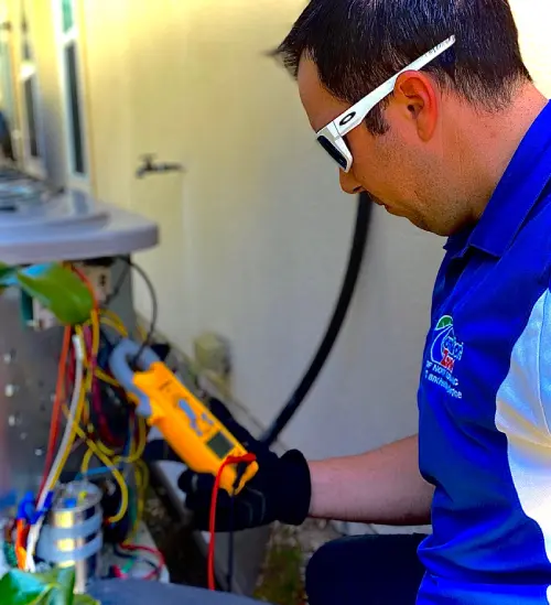 A technician wearing glasses and a blue uniform uses a multimeter to check an outdoor hvac unit.