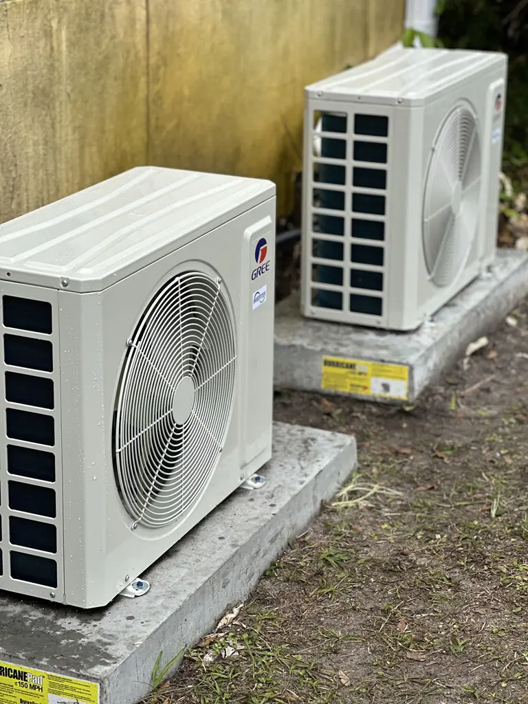 Two gree air conditioning units installed on concrete bases beside a wooden fence.