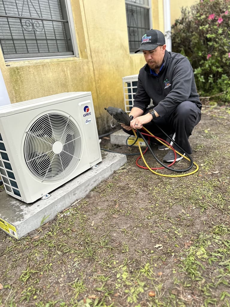 A technician kneels while servicing an outdoor air conditioning unit, holding tools and wearing a cap.