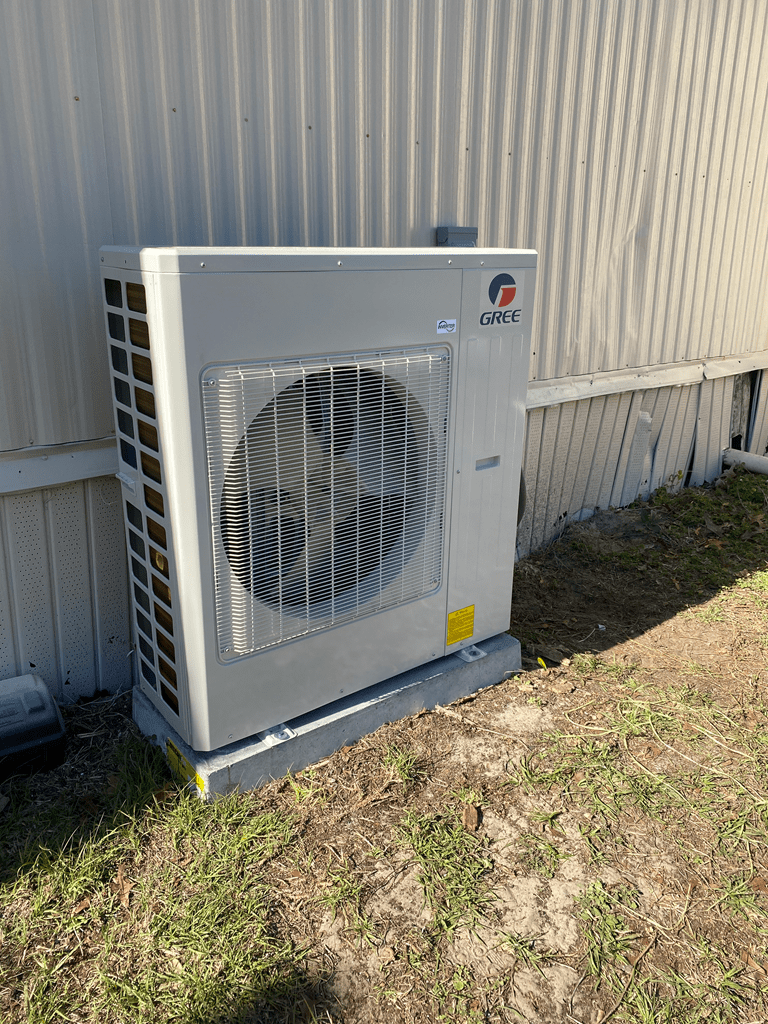 A gree air conditioning unit installed outside next to a metal fence, with visible grass and soil around it.