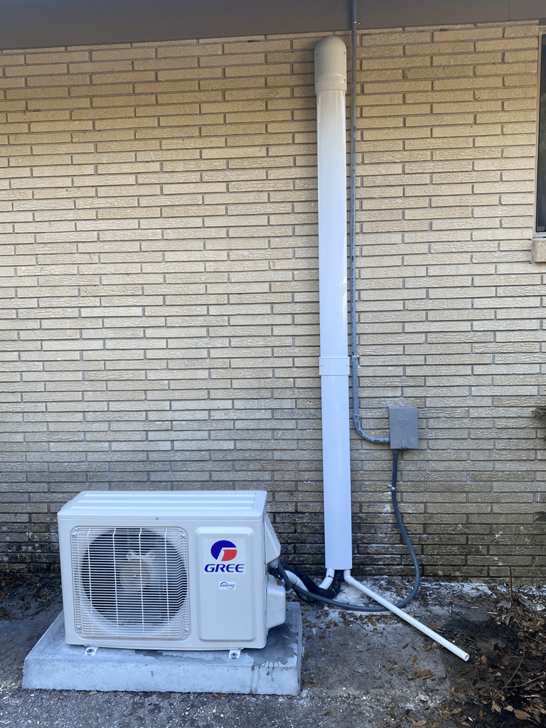 An outdoor hvac unit by a brick wall, connected to a large white vertical pipe and electrical conduit.
