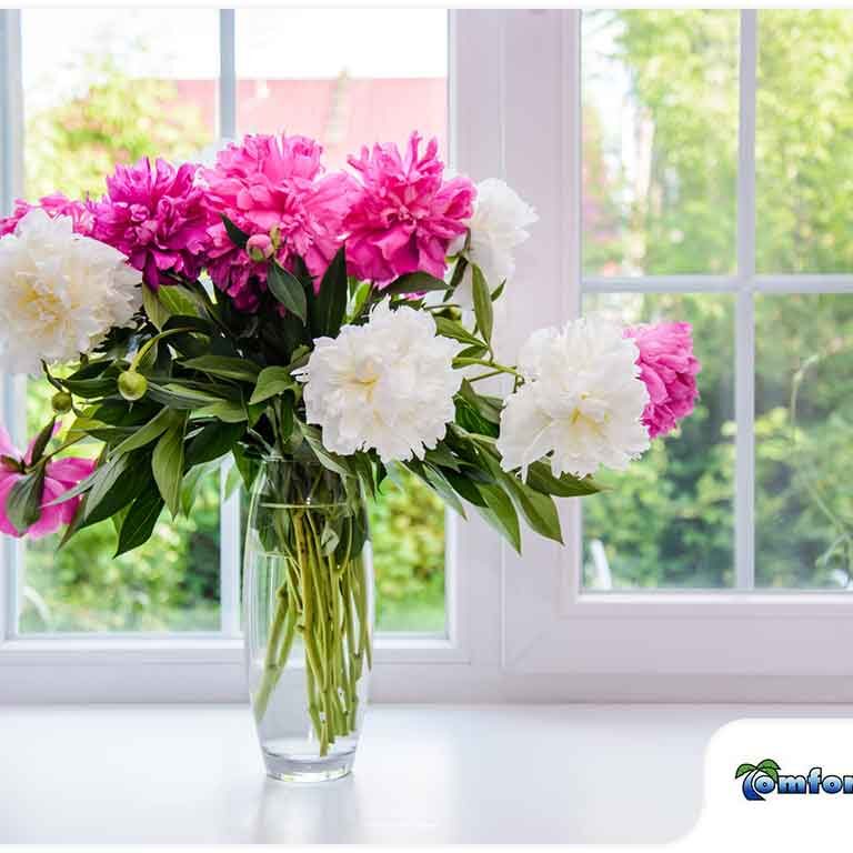 A bouquet of pink and white peonies in a clear vase on a windowsill, with a green garden view through the open window.
