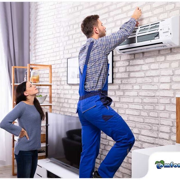 A technician in a blue uniform installs an air conditioner as a woman observes in a modern living room with brick wall.