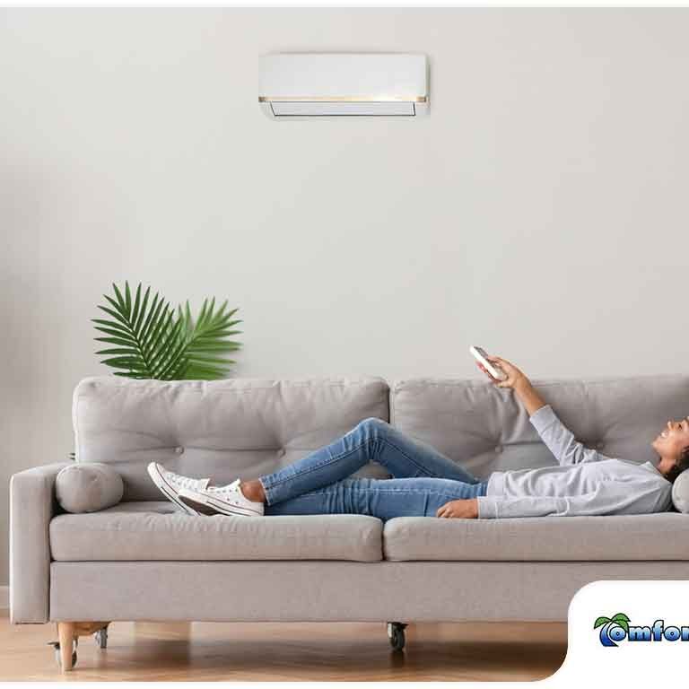 A woman relaxing on a sofa while using a remote control under an air conditioning unit in a modern living room.