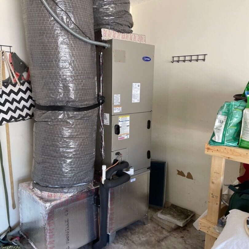 A residential furnace in a cluttered garage, with ductwork, gardening supplies, and household items visible around it.