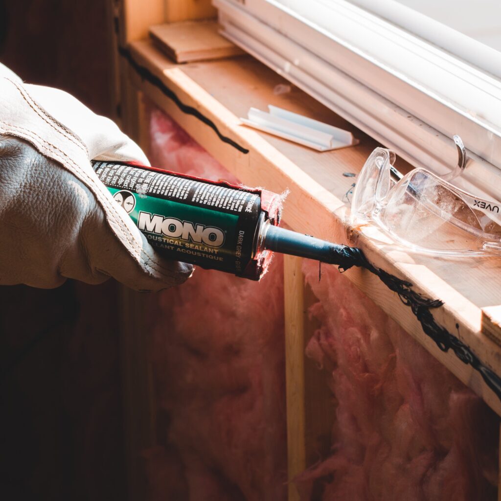 A worker's gloved hand applying sealant from a caulking gun to a window corner in a room under construction.