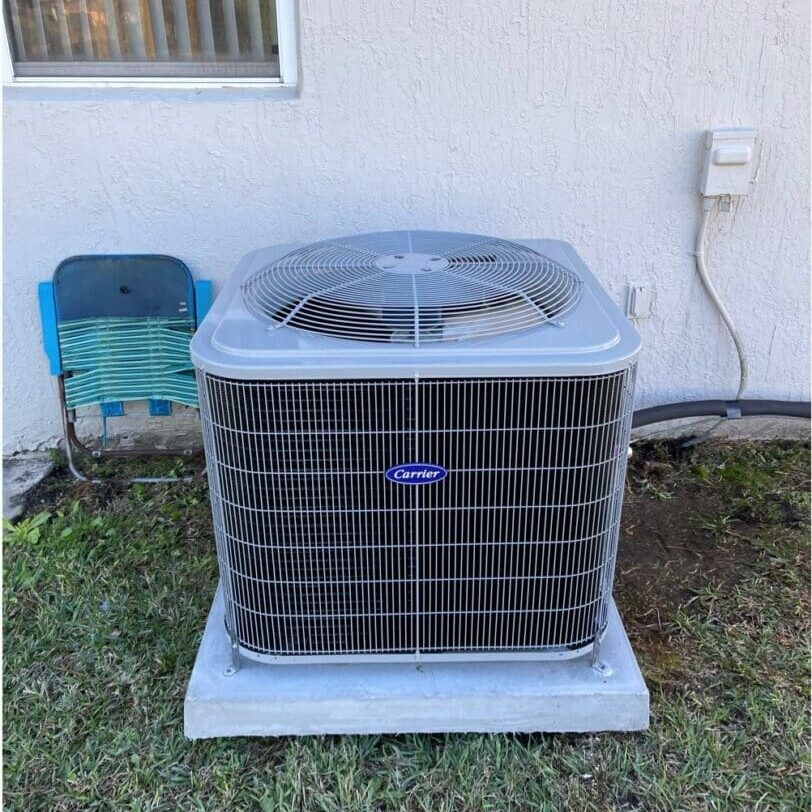 An outdoor hvac unit mounted on a concrete slab next to a building, with a small blue folding chair beside it.