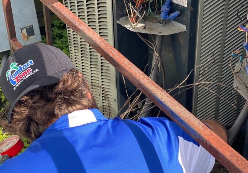 A technician in a blue shirt and cap inspects the internal components of an outdoor hvac unit.
