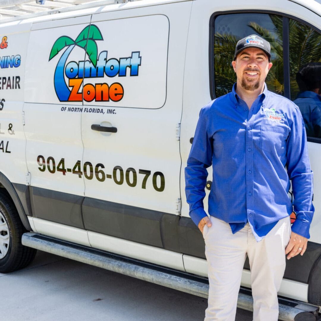 A Comfort Zone of North Florida service technician is standing in front of an air conditioning van, ready to assist with HVAC services, wearing a uniform and a confident smile.