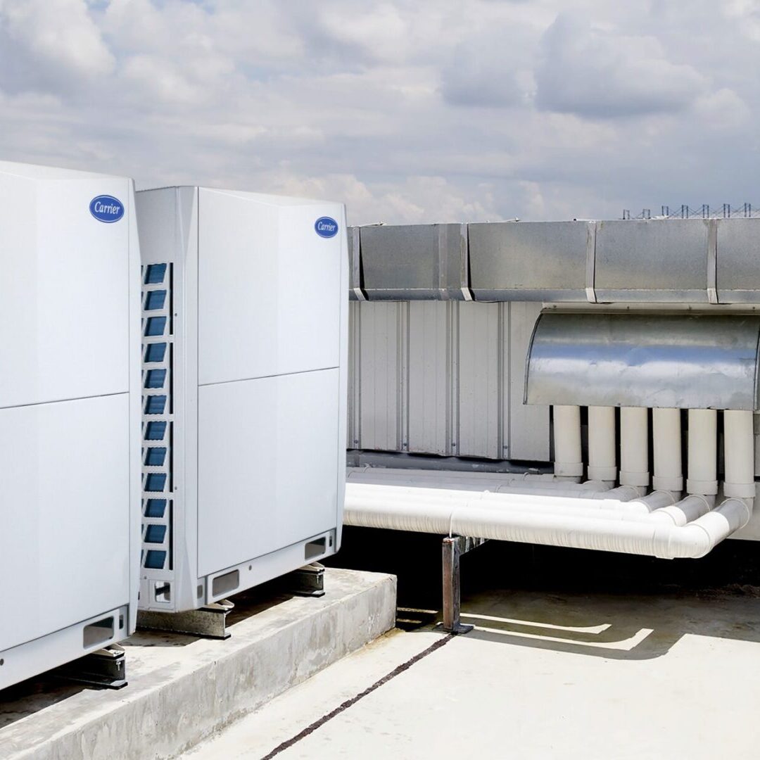 Two large industrial air conditioning units on a building rooftop, connected to ventilation ducts under a cloudy sky.