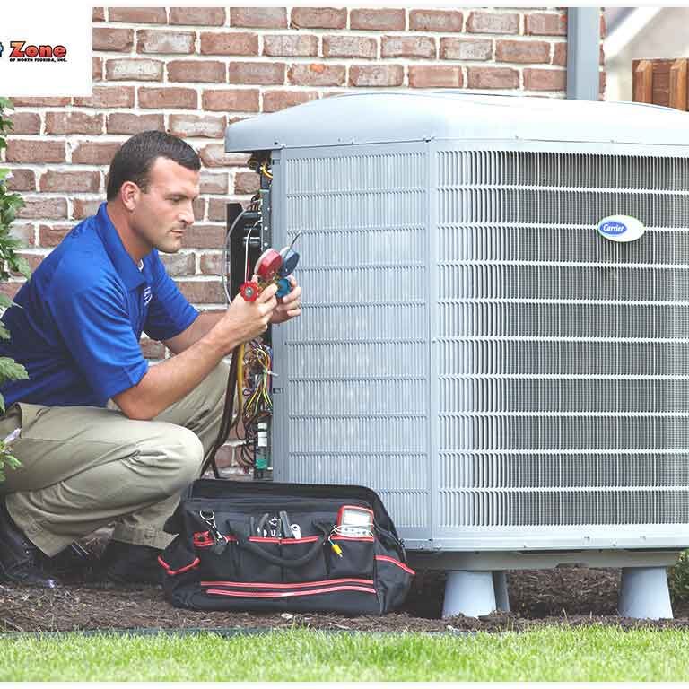 A technician in a blue shirt and beige pants services an outdoor air conditioning unit with tools beside him.