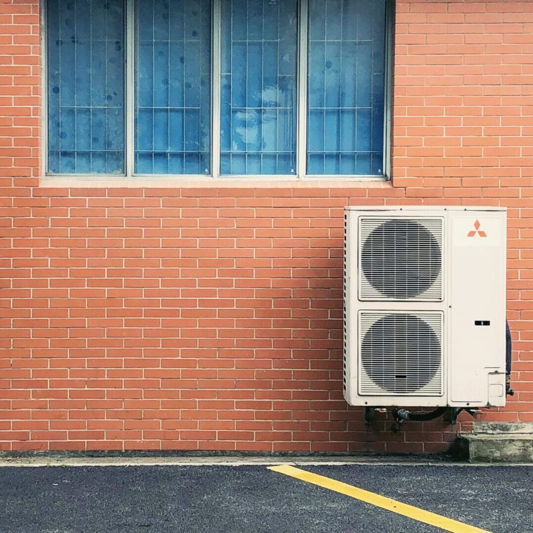An air conditioning unit mounted on a brick wall next to a window with blue shutters, above an asphalt parking lot.