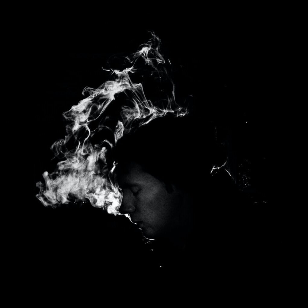 A monochrome image of a person exhaling smoke, creating a swirling pattern against a dark background.