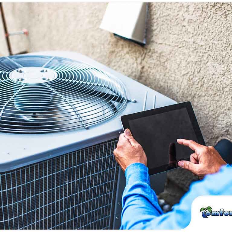 A technician uses a tablet to diagnose or control an air conditioning unit outdoors, with a company logo "comfort zone" visible.