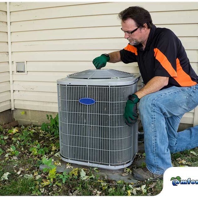 A technician in gloves and sunglasses services an outdoor air conditioning unit beside a house.