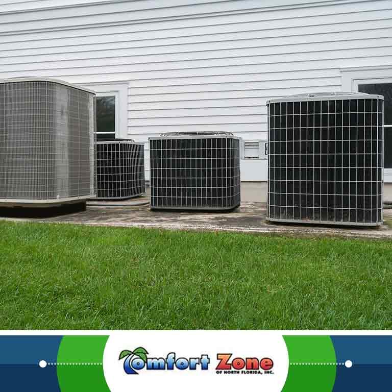 Four air conditioning units of varying sizes installed outside a white-sided building, set on a concrete slab with a grassy backdrop.