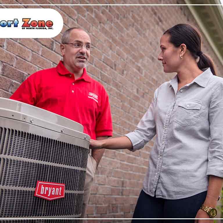 A woman listens attentively to a male technician in a red shirt discussing an outdoor air conditioning unit by a brick house.